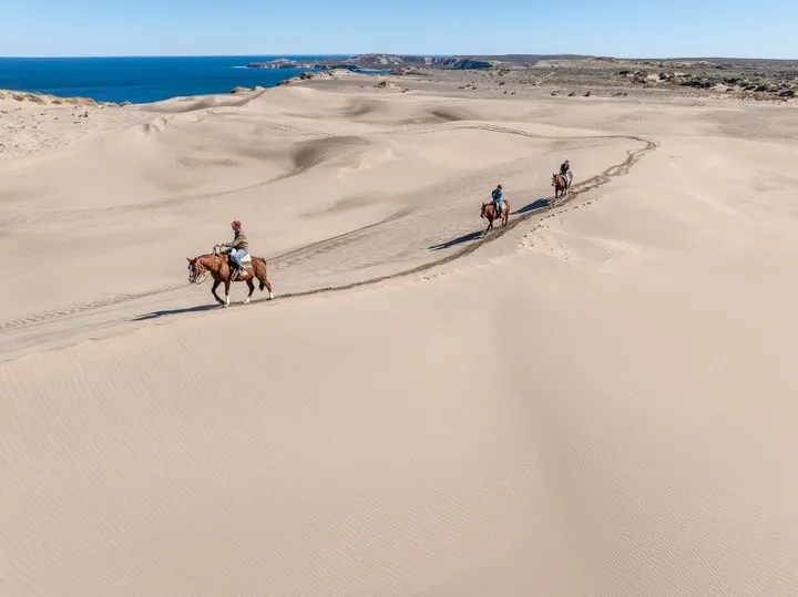 Vista aérea de tres jinetes cruzando los médanos con el mar de fondo